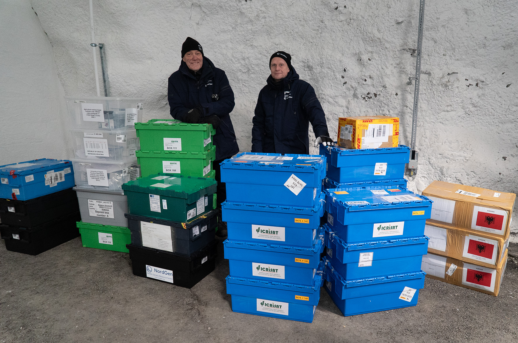 Two men wearing warm clothes standing behind piles of boxes in a storage room. 