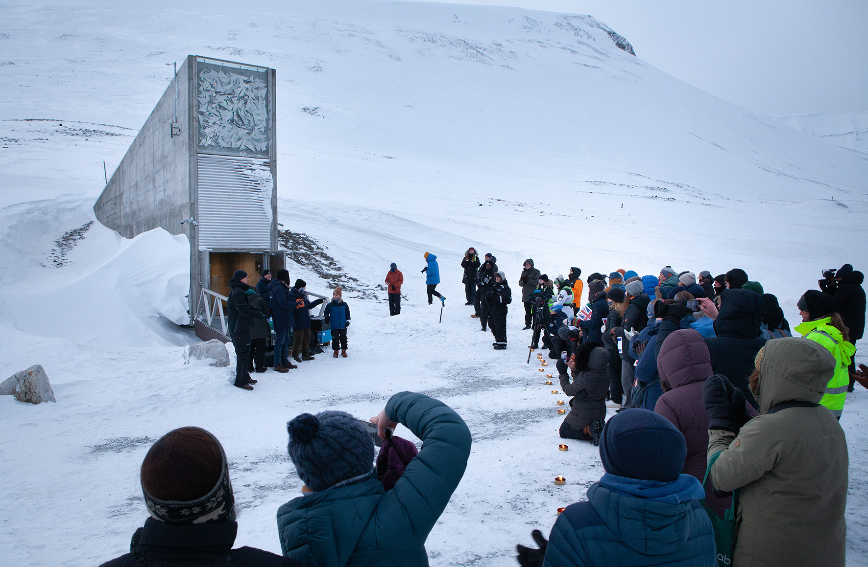 A large group of people standing outdoors infront of a wedge-shaped building, snow is on the ground. 